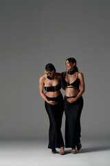 Happy, beautiful pregnant twins sisters in black fashionable and elegant clothes posing together, smiling and touching their bellies. Young women expecting babies.