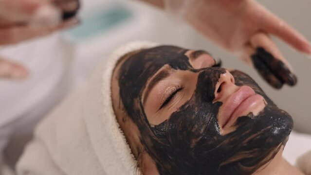 Mud therapy, Anti-acne care, Mineral mask. Beauty salon, skincare professional applies black mask to patient's face, promoting detoxification, lifting, kelp rejuvenation, mud therapy, anti-acne