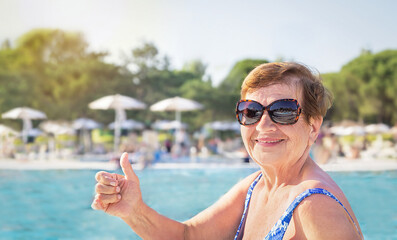 Active senior (elderly) woman (over age of 50) in swimsuit and sunglasses showing thumbs up on...