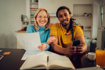 A happy interracial couple is sitting together at home and calculating bills and taxes on the phone.