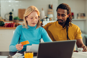 An interracial serious couple is sitting at the dining table at home and using a credit card for...