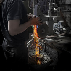 Man in a dark forge grinding a knife with a belt grinder and sparks flying downwards