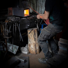 Man in a dark forge workshop heating red hot steel with a gas blast furnace