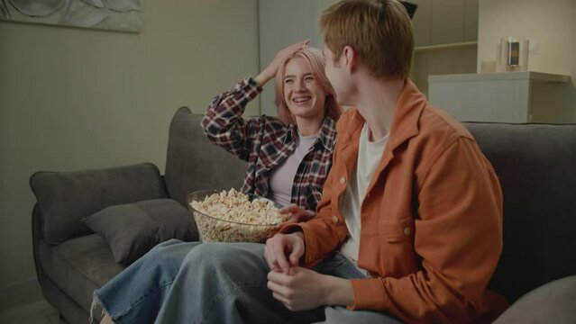 Millennial Couple Having Fun on Couch. Young adult man and woman eating popcorn and laughing.