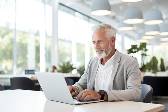 An older attractive man with white hair and a beard is sitting in the office at a laptop, generative ai