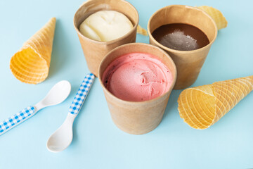 Ice cream in a craft box with different flavors - strawberry, chocolate, vanilla stands on a blue background along with a waffle cone and a checkered spoon. View from above.