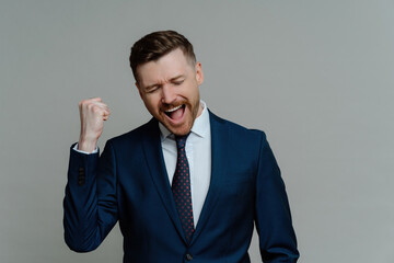 Successful businessman celebrates, clenches fist, exclaims in joy, dressed formally, satisfied after deal. Grey background. Congrats, I did it!