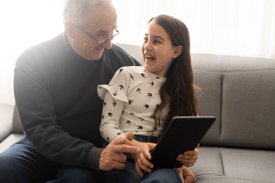 Cute little girl and her handsome grandpa are smiling while sitting on couch at home. Girl is using a tablet. - Powered by Adobe