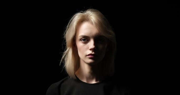 A serious Caucasian woman standing against a striking black background. With an unwavering gaze directed towards the camera, she exudes a profound sense of seriousness and determination.