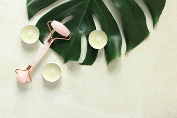 Facial massage roller with two rose quartz heads. Facial beauty massage, spa self-care concept. Massage roller on a monstera leaf, candles, top view