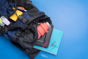 Back to school concept with blue and black book bag backpack, contents spilling out. Spiral notebook, tablet, water bottle, paper clips, note pads, pencils, pens, and highlighters.