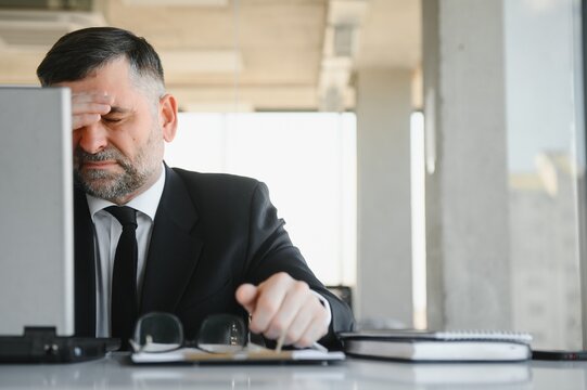 Worried businessman in dark suit sitting at office desk full with books and papers being overloaded with work.