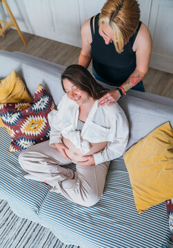 Young woman tender massaging pregnant partner's female shoulders. Same-sex marriage couple on home living room sofa. Woman's health, happy pregnancy doula supporting and calm mental mood concept image
