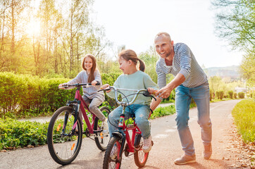 Smiling father with two daughters during outdoor walk. He teaching younger girl to ride a bicycle....