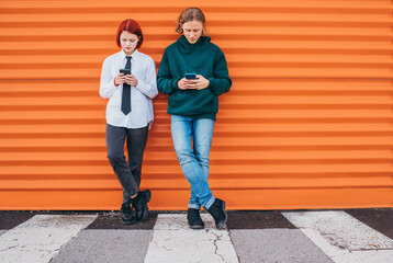 Fototapeta na wymiar ..Two caucasian teen friends boy and a girl browsing their smartphone devices while they lean on the orange wall background. Careless young teenhood . time and a modern technology concept image.