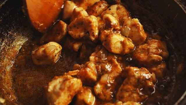Chicken pieces in spices fried in a cast-iron skillet are mixed with a wooden spatula