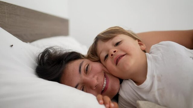 Mother and son laid in bed in embrace looking at camera smiling. Motherhood lifestyle child and mom together
