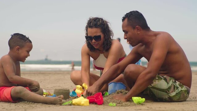 happy couple making sand castle at tropical beach with their son. Child with mom and dad outdoor at seaside playing together
