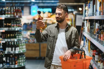 Fototapeten A man takes alcoholic drinks from the supermarket shelf. Shopping for alcohol in the store © Serhii