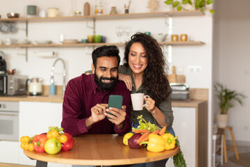 Happy arab couple using smartphone, ordering food delivery or shopping online in kitchen interior