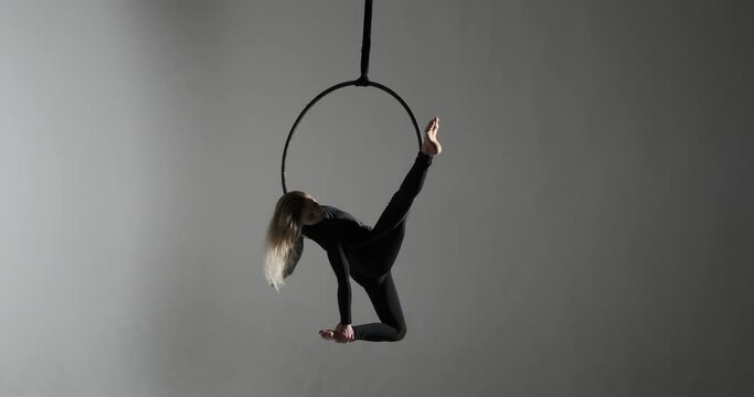 In a studio illuminated by contrasting lights against a dark background, a caucasian woman showcases her acrobatic skills on an aerial hoop. With her long hair cascading down.
