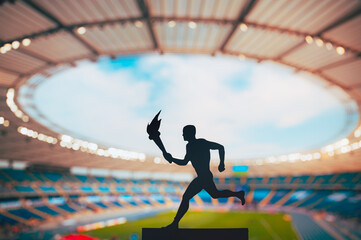 Silhouette of male athlete holding torch relay. Modern Track and Field Stadium in background. Photo for Summer game 2024 in Paris