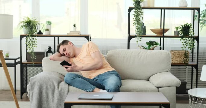 A sleepy and bored Caucasian man sits on a comfortable sofa in a cozy living room. He has a phone in his hand, which he uses to check the time. He suffers from boredom.