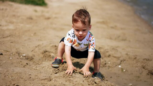 Sweet Caucasian kid claps his hands dirty from sand. Toddler boy steps and sits to the ground deepening hands into sand again. Sunny day backdrop.
