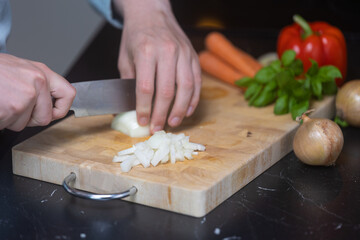 Man in blue shirt cutting and dicing onion og wooden cutting board, with other vegetables in the background.