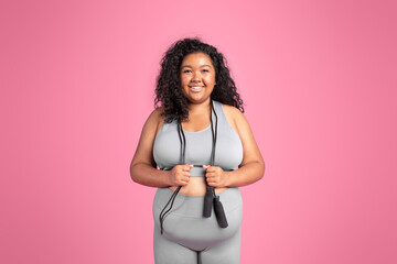 Excited black overweight woman in sportswear holding jump rope and smiling at camera on pink studio...