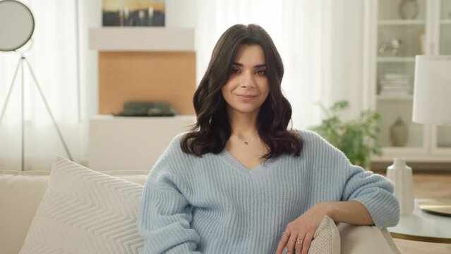 Positive relaxed hispanic woman posing in front of decorative fireplace in modern living room. Slow motion people 4K. Smiling beautiful latin woman looking at camera sitting on sofa alone at home