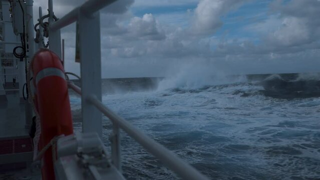 High waves and splashes along side. Ship in storm. White foam on sea. Deck railing and lifebuoy. Strong pitching. Vessel climb up wave.
