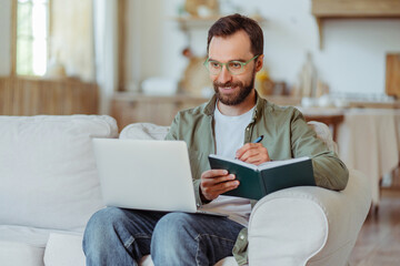 Fototapeta premium Handsome smiling bearded businessman wearing eyeglasses using laptop computer, taking notes, watching training courses at home. Smart student studying, learning language, online education concept
