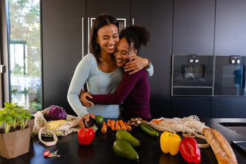 Happy biracial lesbian couple hugging and unpacking groceries in kitchen