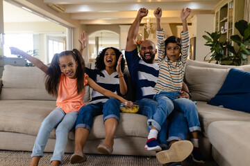 Happy biracial family sitting on couch with snacks, watching tv and cheering