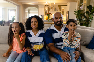 Focussed biracial family sitting on couch eating snacks and watching tv