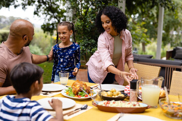 Happy biracial parents, son and daughter serving food and sitting at table for meal on terrace