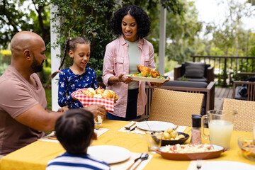 Happy biracial parents, son and daughter serving food and sitting at table for meal on terrace