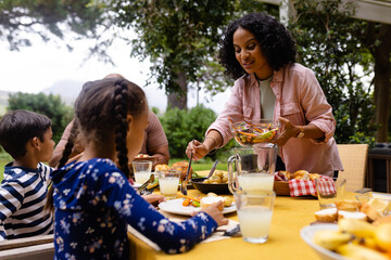 Happy biracial parents, son and daughter serving food and sitting at table for meal in garden