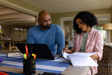 Serious biracial parents using laptop, looking at bills sitting at table with daughter in background