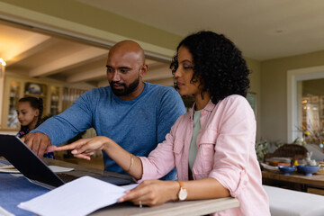 Biracial parents using laptop, looking at bills and talking at table with daughter in background