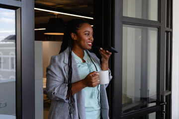 Plus size african american casual businesswoman with cup of coffee talking on smartphone in office