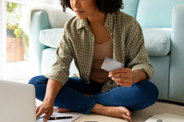 Mid section of biracial woman using laptop and credit card sitting on floor at home - Powered by Adobe