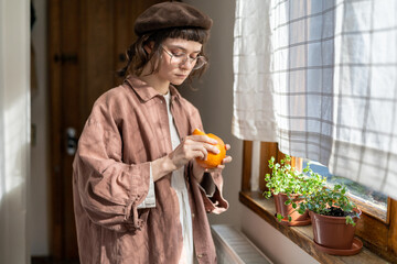 vitamin C deficiency. Millennial hipster girl peeling citrus fruit while standing by window in...