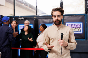 Male TV reporter standing in shopping mall during Black Friday sales, making news for television. Young man journalist holding microphone talking to camera while standing against crowd of shoppers