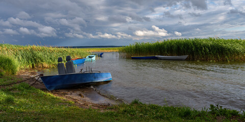 fishing boats parked in a small lake bay overgrown with reeds under a cloudy sky in windy weather before rain