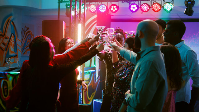 Smiling people clinking glasses filled with alcohol on dance floor, saying cheers under colorful spotlights. Young men and women enjoying cool funky party, entertainment. Handheld shot.