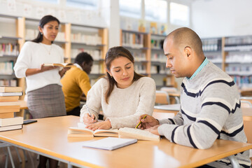 Fototapeta premium Young adult people studying together in library, talking, reading books and writing in notepads