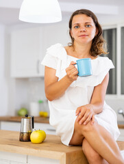 Fototapeta na wymiar Cheerful woman drinking fresh coffee in kitchen at home. Woman sitting on table.