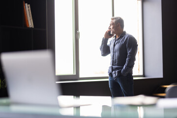 Smiling mature business man talking on cell phone using laptop in modern office.